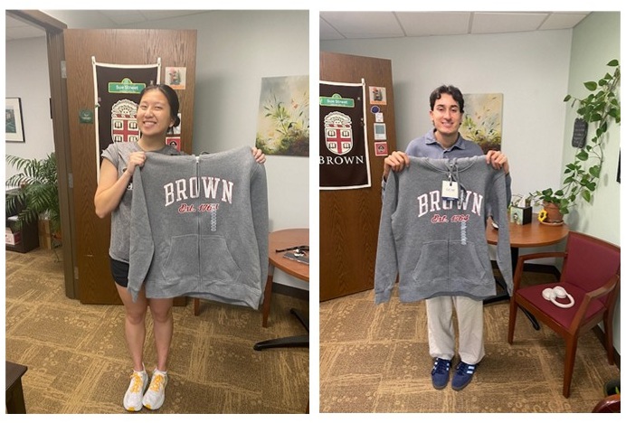 Two sophomore students , each holding a Brown University grey zip-up hoodie.