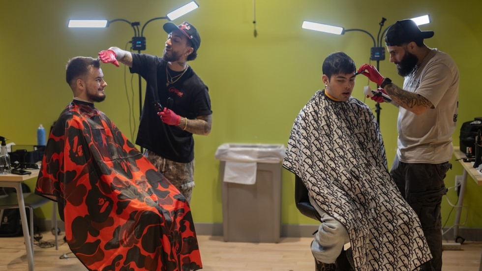 2 students sitting in chairs having their hair cut by 2 barbers.