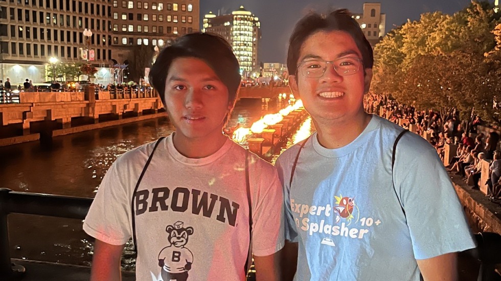 Two students standing on the bridge over the Providence river, a row of small fires is lit on the river behind them.