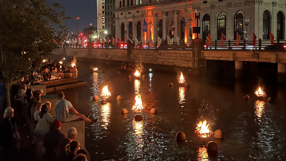 Providence riverfront with fires lit in the river and many people standing around gazing at it.